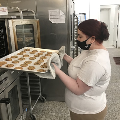 Photo of Jolie, a Sunflower Bakery employee putting tray of cookies into oven in professional kitchen.